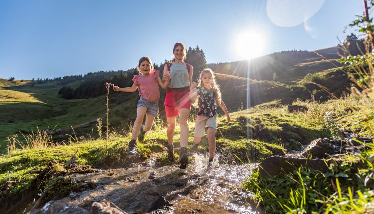Eine Frau und zwei Mädchen wandern über einen kleinen Bach in einer sonnigen, grünen Berglandschaft.