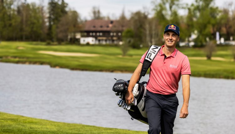 Ein lächelnder Golfer in einem rosa Hemd trägt eine Golftasche neben einem See auf einem üppigen, grünen Golfplatz.