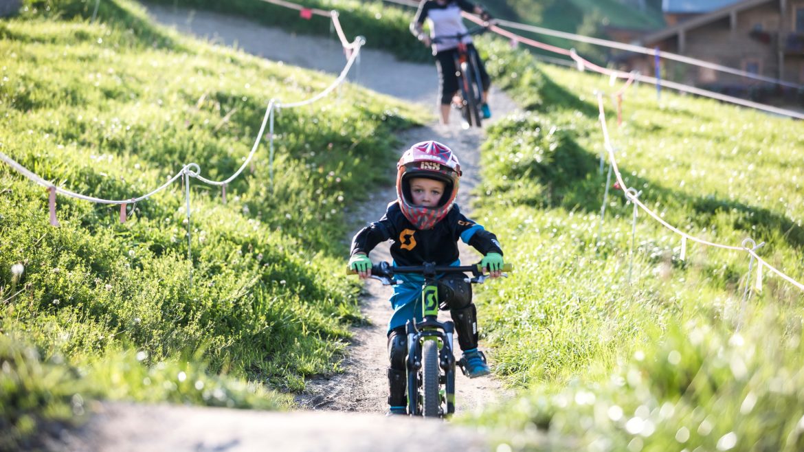 Ein junges Kind mit Helm fährt mit dem Fahrrad auf einem grasbewachsenen Weg, im Hintergrund ein erwachsener Radfahrer.
