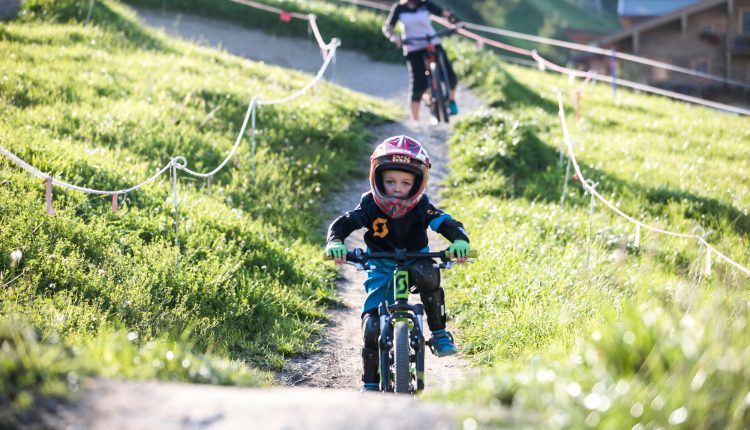 Ein junges Kind mit Helm fährt mit dem Fahrrad auf einem grasbewachsenen Weg, im Hintergrund ein erwachsener Radfahrer.