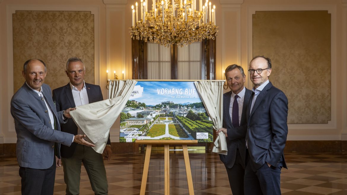 Vier Männer enthüllen in einem eleganten Raum mit Kronleuchtern und Marmorböden ein Poster mit Architekturplänen.