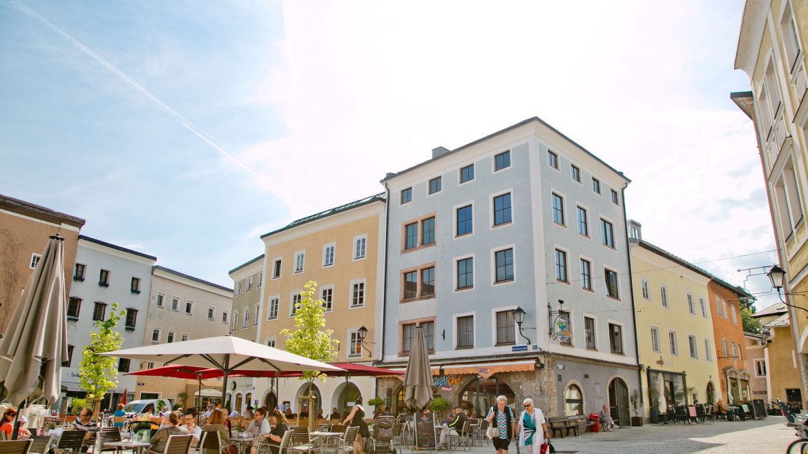 Ein Straßencafé auf einem europäischen Platz mit Menschen, die dort essen, Sonnenschirmen und pastellfarbenen Gebäuden an einem sonnigen Tag.