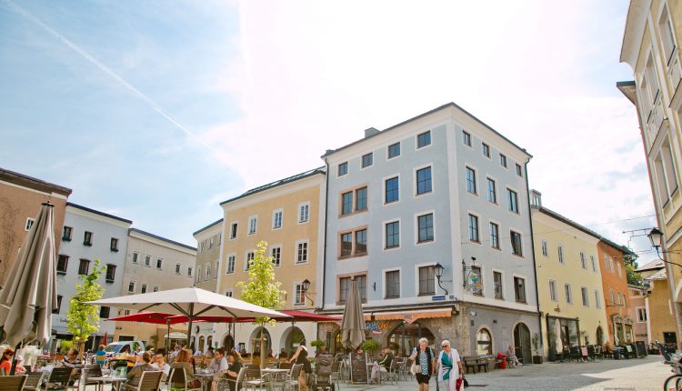 Ein Straßencafé auf einem europäischen Platz mit Menschen, die dort essen, Sonnenschirmen und pastellfarbenen Gebäuden an einem sonnigen Tag.