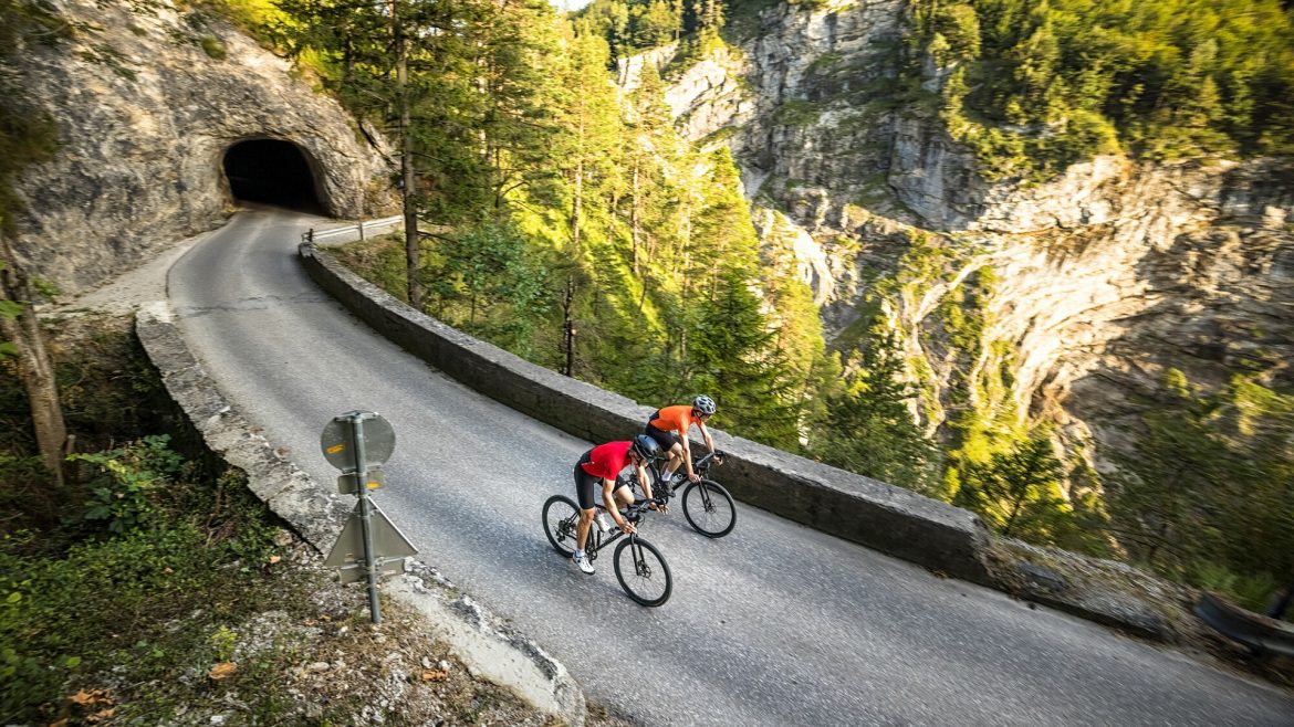 Zwei Radfahrer fahren auf einer kurvenreichen Bergstraße in der Nähe eines Tunnels, umgeben von Bäumen und felsigen Klippen.