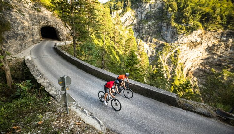 Zwei Radfahrer fahren auf einer kurvenreichen Bergstraße in der Nähe eines Tunnels, umgeben von Bäumen und felsigen Klippen.