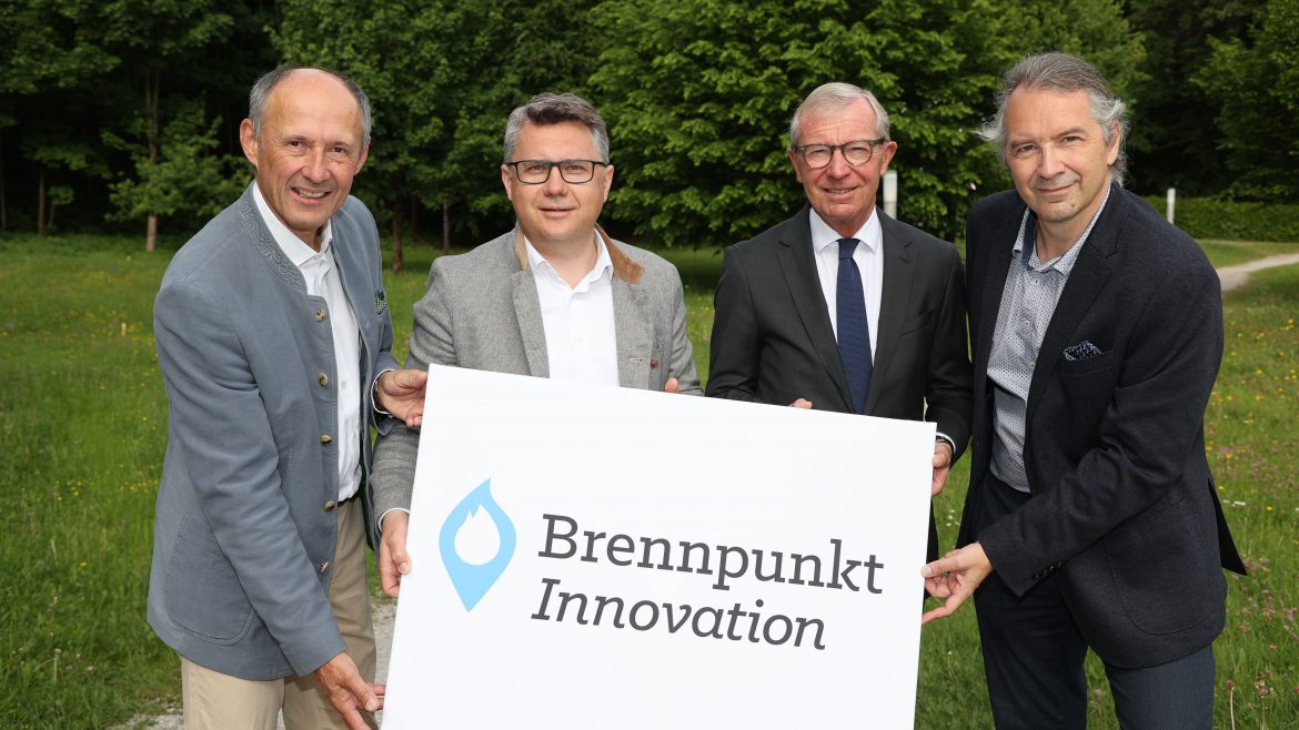 Vier Männer stehen im Freien und halten ein Schild mit der Aufschrift Brennpunkt Innovation und einem blauen Flammenlogo.