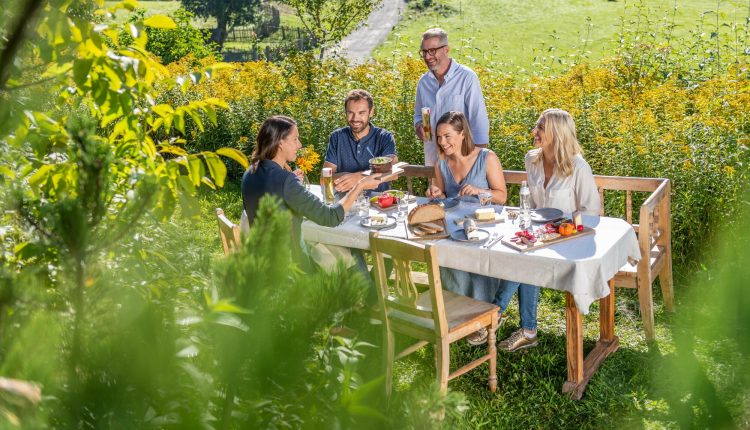 Fünf Personen, die an einem sonnigen Tag an einem Tisch im Freien in einem üppigen, grünen Garten eine Mahlzeit genießen.