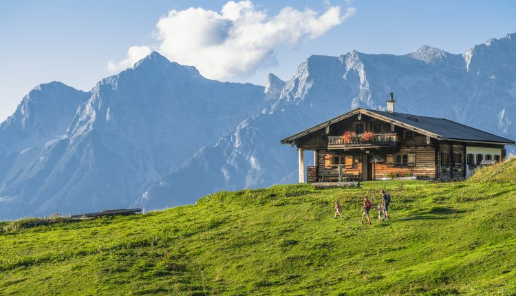Holzhütte auf einem grünen Hügel mit spazierenden Menschen vor hohen, felsigen Bergen und einem blauen Himmel.