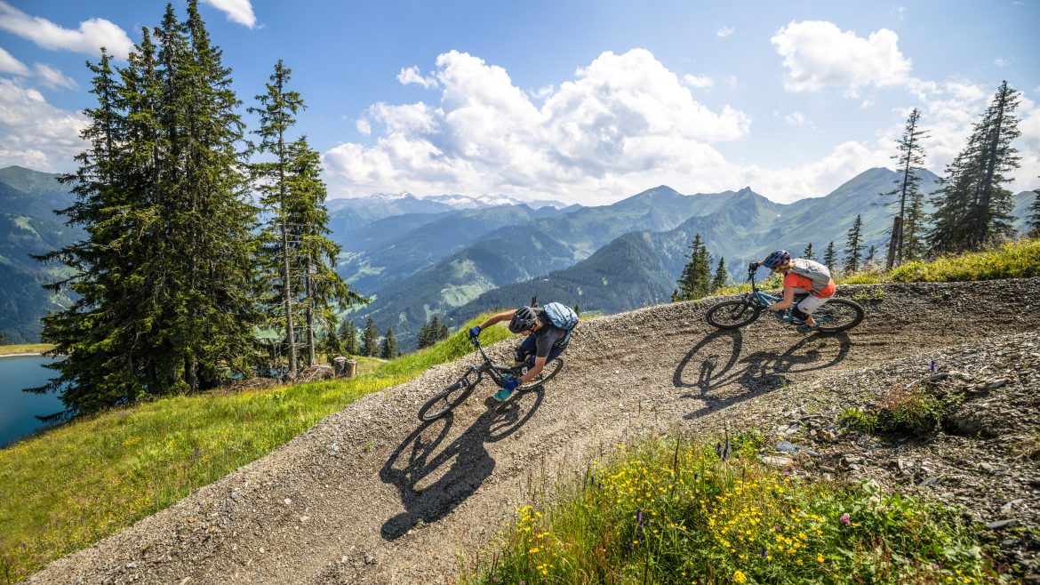 Zwei Radfahrer fahren mit ihren Mountainbikes auf einer kurvenreichen Strecke mit Bergen und Bäumen unter einem sonnigen Himmel.