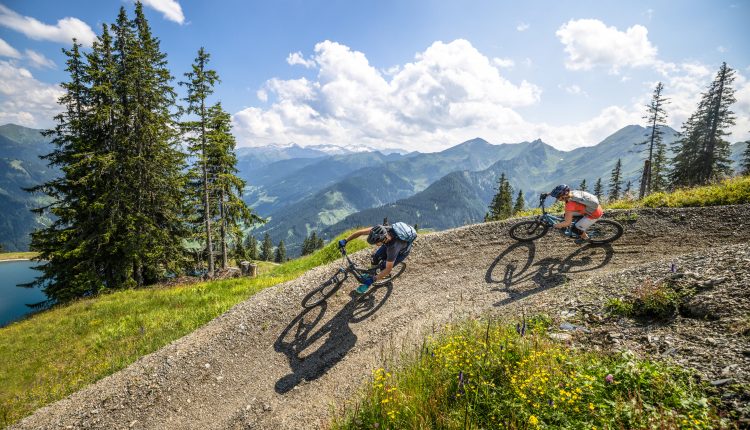 Zwei Radfahrer fahren mit ihren Mountainbikes auf einer kurvenreichen Strecke mit Bergen und Bäumen unter einem sonnigen Himmel.
