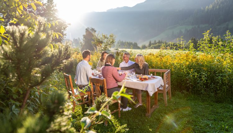 Fünf Personen genießen eine Mahlzeit im Freien an einem Tisch in einem üppigen, sonnenbeschienenen Garten mit Bergen im Hintergrund.
