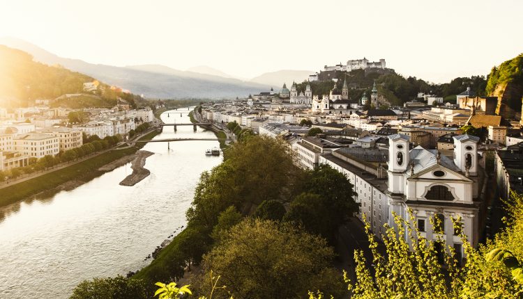 Eine malerische Ansicht von Salzburg, Österreich, mit einem Fluss, Brücken und historischen Gebäuden bei Sonnenuntergang.