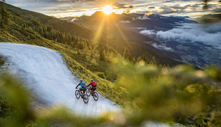 Zwei Radfahrer fahren bei Sonnenuntergang eine kurvenreiche Bergstraße hinauf, umgeben von üppigem Grün und fernen Wolken.