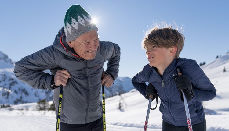 Ein älterer Mann und ein Junge lächeln sich beim Skilanglauf auf einem sonnigen, verschneiten Berg an.