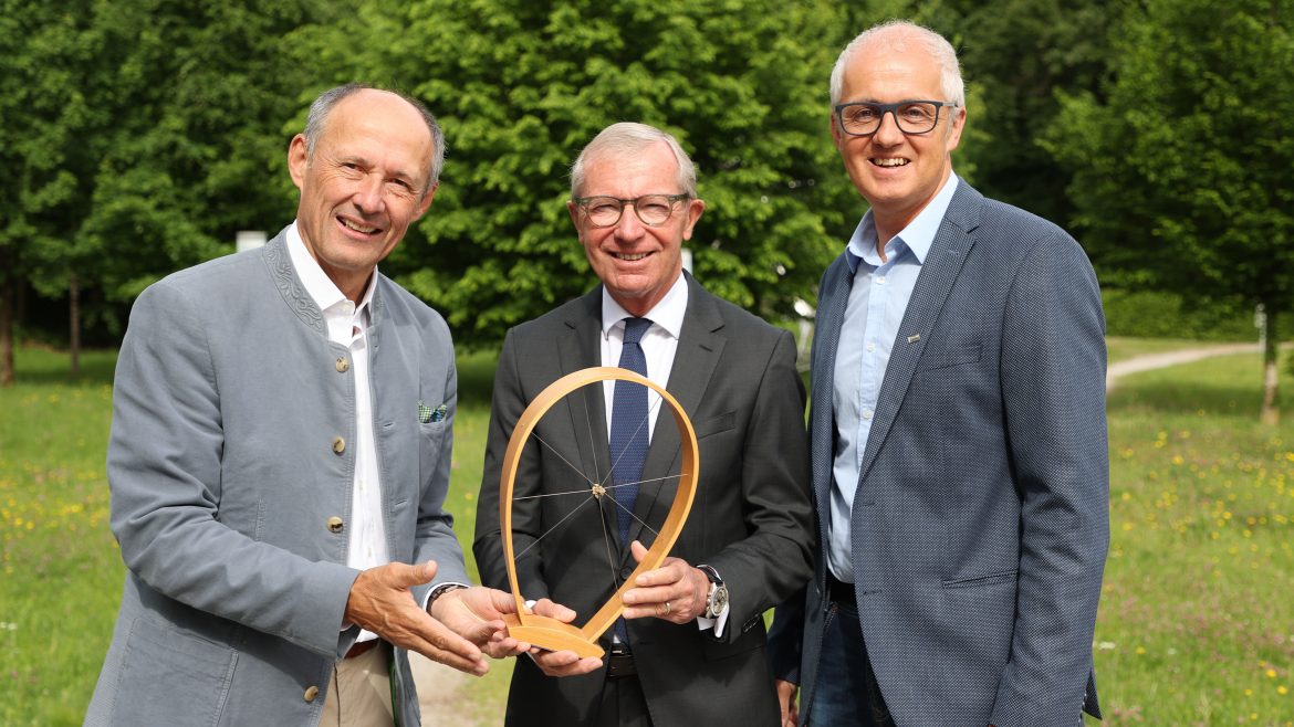 Drei Männer in Anzügen stehen im Freien, halten einen hölzernen Preis mit einem Draht in der Mitte und lächeln in die Kamera.