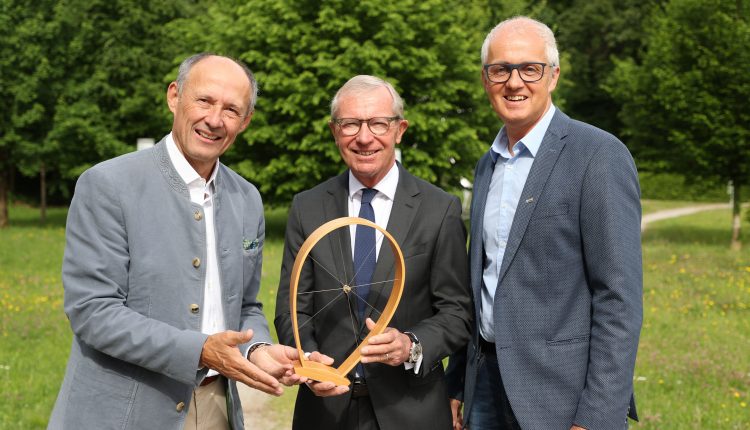 Drei Männer in Anzügen stehen im Freien, halten einen hölzernen Preis mit einem Draht in der Mitte und lächeln in die Kamera.