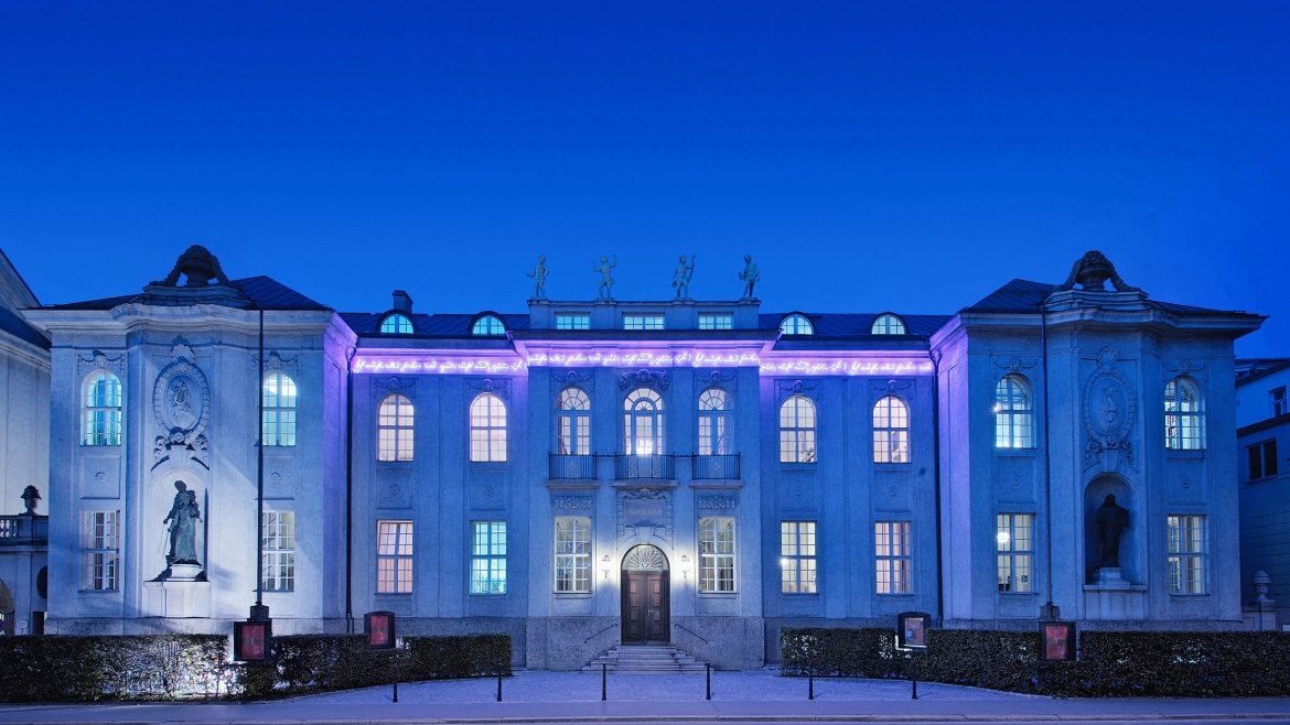 Elegantes Gebäude mit violetter Beleuchtung und Statuen, fotografiert in der Abenddämmerung vor einem tiefblauen Himmel.