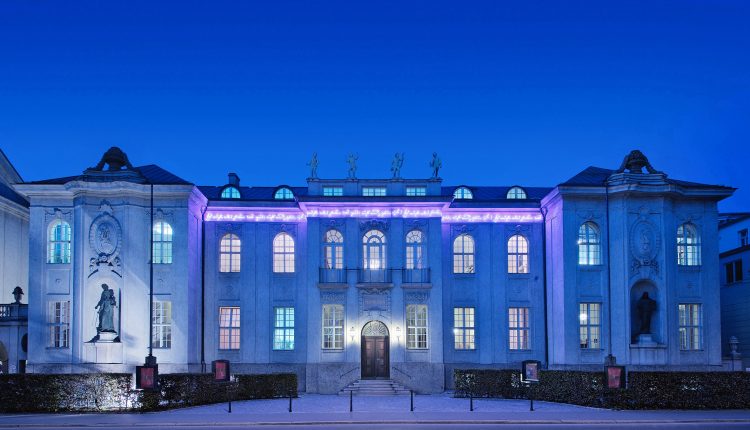 Elegantes Gebäude mit violetter Beleuchtung und Statuen, fotografiert in der Abenddämmerung vor einem tiefblauen Himmel.