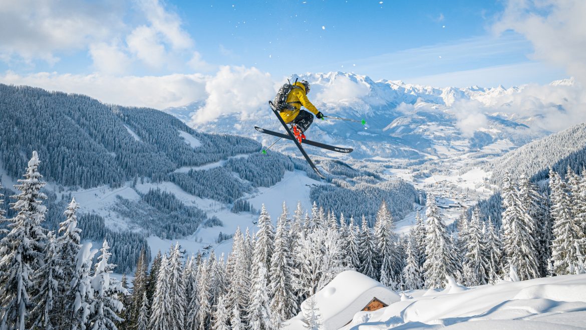 Ein Skifahrer in einer gelben Jacke springt mitten in der Luft über eine verschneite Berglandschaft mit Kiefern.