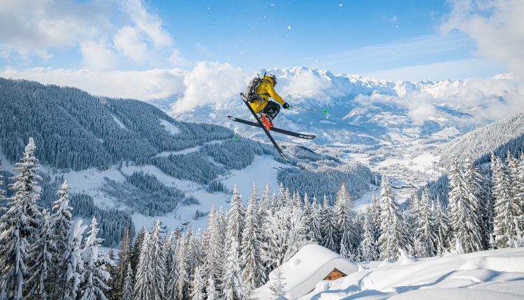 Ein Skifahrer in einer gelben Jacke springt mitten in der Luft über eine verschneite Berglandschaft mit Kiefern.
