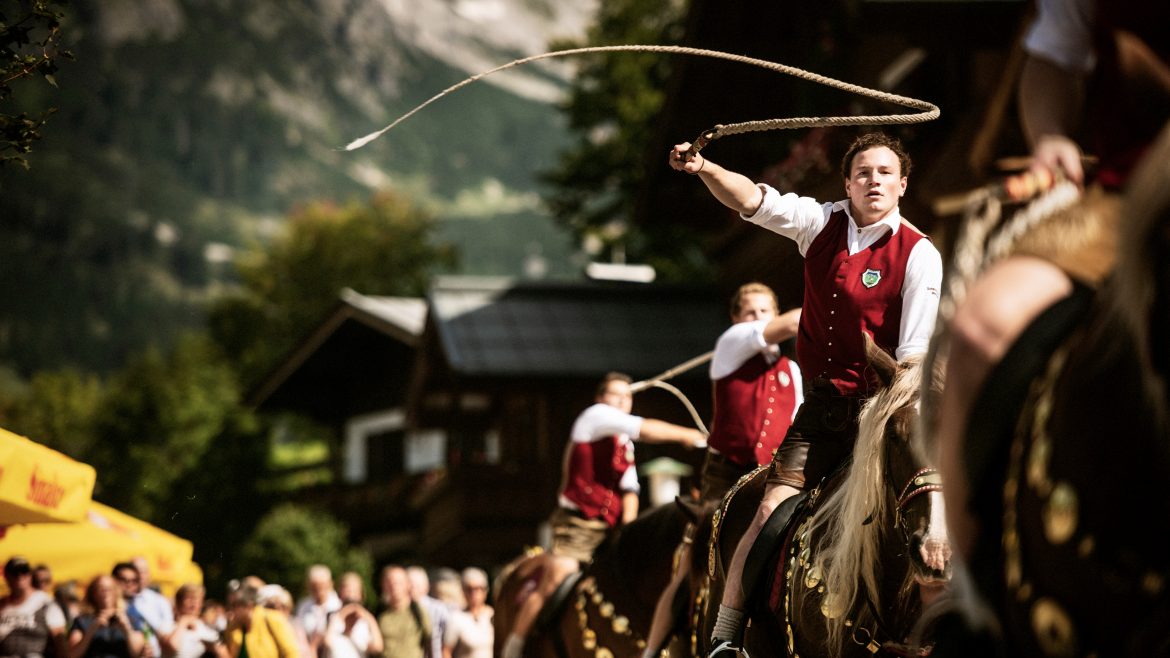 Ein Mann in traditioneller österreichischer Kleidung auf einem Pferd knallt die Peitsche während eines Festes mit einer Menschenmenge und Bergen im Hintergrund.