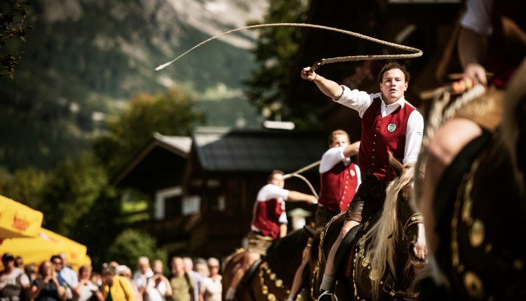 Ein Mann in traditioneller österreichischer Kleidung auf einem Pferd knallt die Peitsche während eines Festes mit einer Menschenmenge und Bergen im Hintergrund.