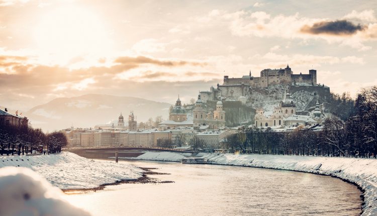 Verschneites Stadtbild von Salzburg mit einem Fluss, Kuppelkirchen und einer Festung auf dem Hügel bei Sonnenuntergang.