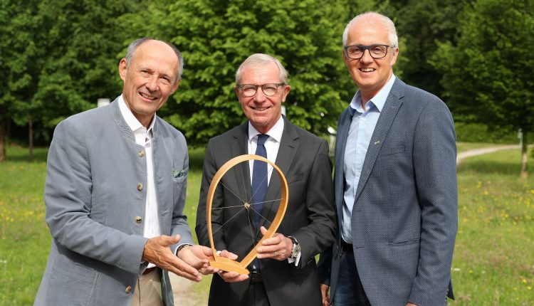 Drei lächelnde Männer in Anzügen stehen im Freien und halten einen hölzernen Preis mit einem radähnlichen Mittelstück.