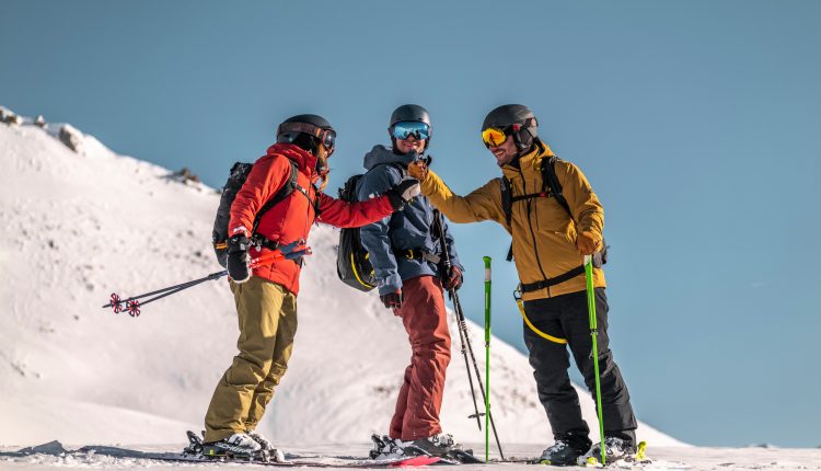 Drei Skifahrer in bunten Klamotten unterhalten sich und lachen auf einem verschneiten Berg unter einem klaren blauen Himmel.