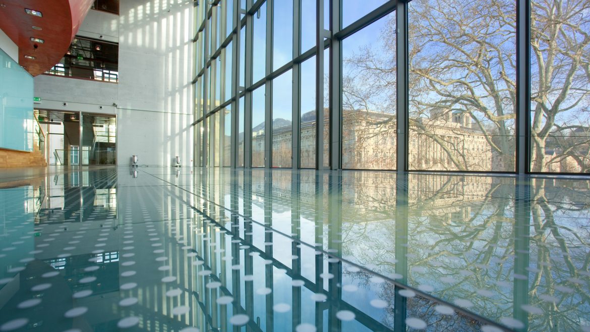 Modernes Gebäude mit Glaswänden und Spiegelungen von Bäumen und Himmel auf dem polierten Boden.