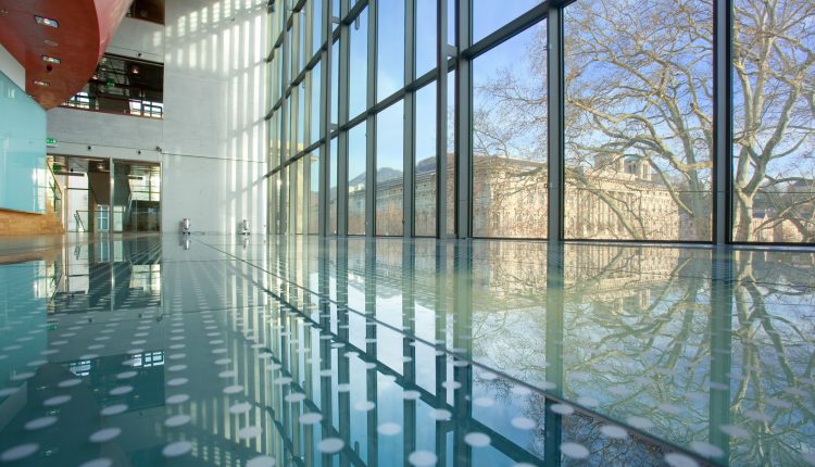 Modernes Gebäude mit Glaswänden und Spiegelungen von Bäumen und Himmel auf dem polierten Boden.