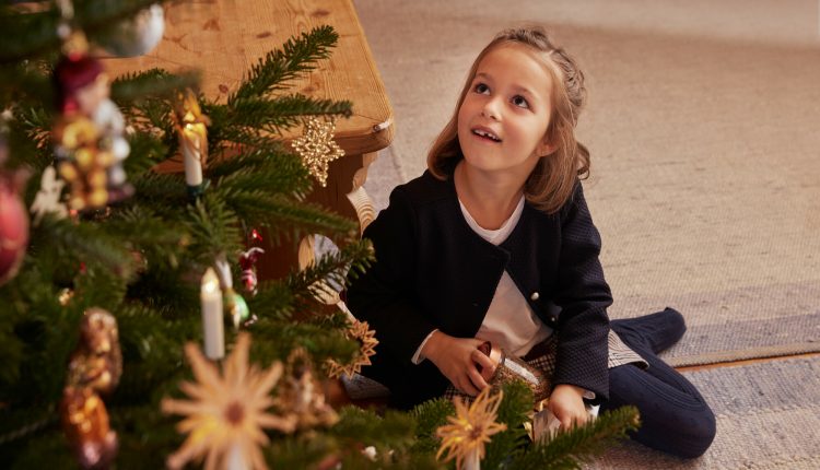 Ein junges Mädchen sitzt an einem geschmückten Weihnachtsbaum, lächelt und schaut aufgeregt nach oben.