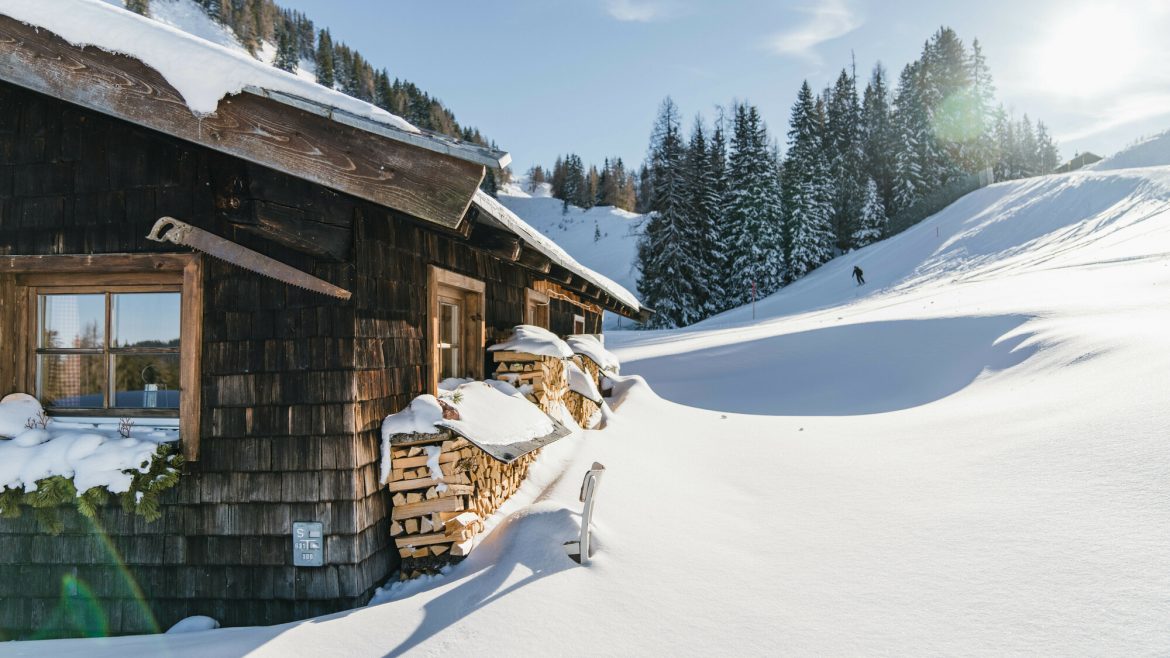 Eine Holzhütte inmitten von Tiefschnee und Kiefern, mit Sonnenlicht und einer Person, die in der Ferne Ski fährt.