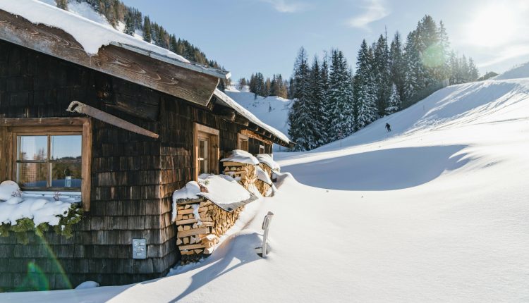 Eine Holzhütte inmitten von Tiefschnee und Kiefern, mit Sonnenlicht und einer Person, die in der Ferne Ski fährt.