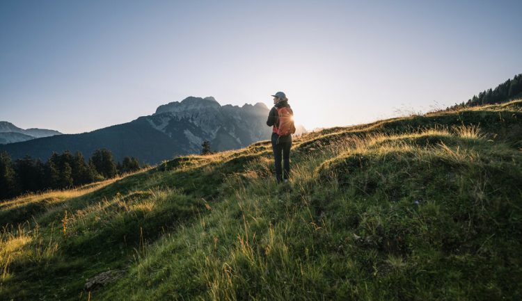 Ein Wanderer steht bei Sonnenaufgang auf einem grasbewachsenen Hügel, mit den Bergen im Hintergrund.