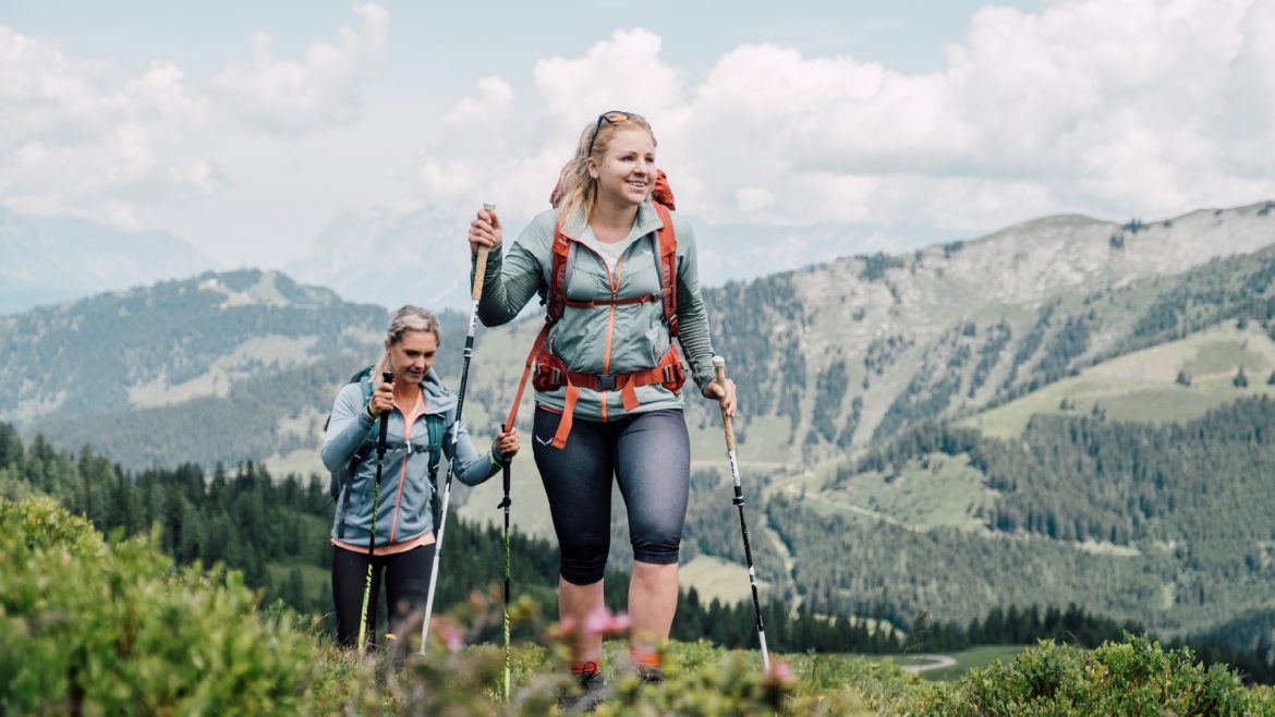 Zwei Frauen wandern mit Trekkingstöcken auf einem grasbewachsenen Bergpfad, mit Hügeln und Wolken im Hintergrund.