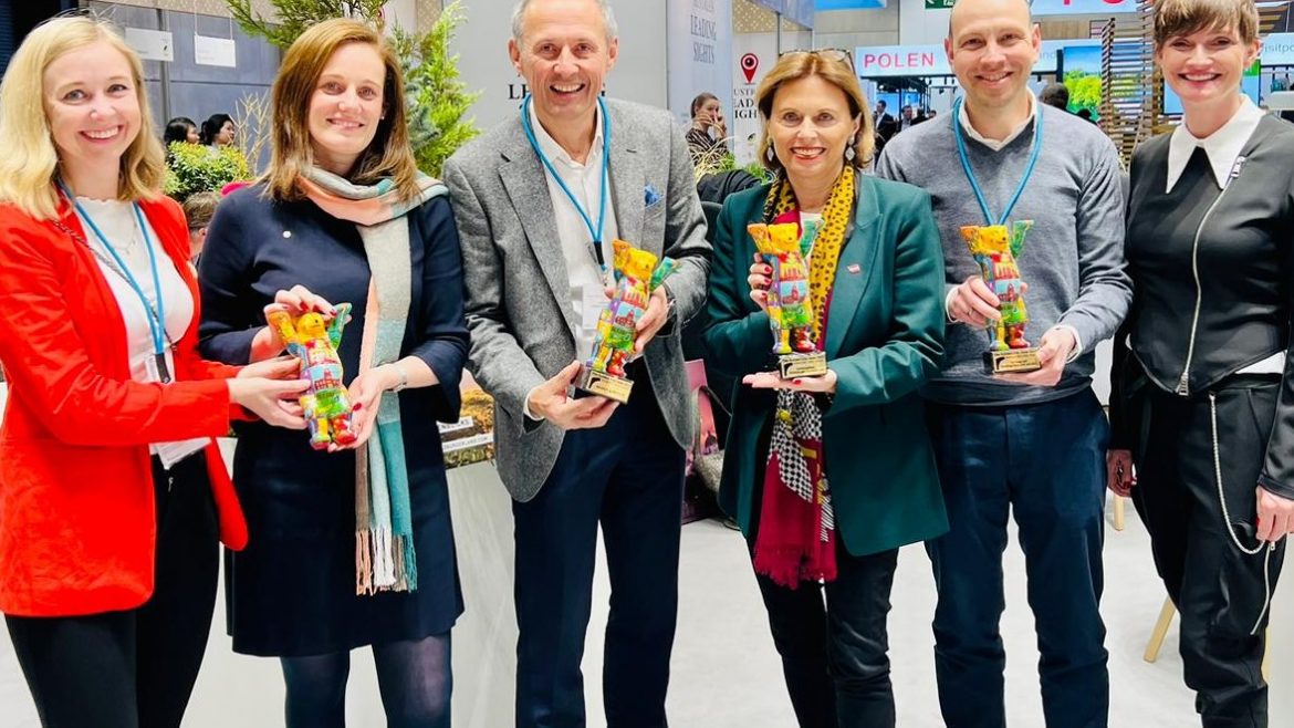 Sechs lächelnde Menschen, die bei einer Veranstaltung oder Ausstellung in einem Raum stehen und jeweils einen bunten, bärenförmigen Preis in der Hand halten.