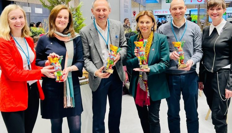 Sechs lächelnde Menschen, die bei einer Veranstaltung oder Ausstellung in einem Raum stehen und jeweils einen bunten, bärenförmigen Preis in der Hand halten.