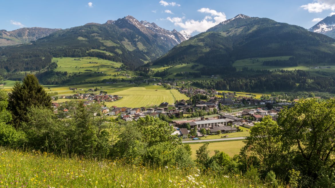 Ein kleines Dorf mit Feldern und Häusern liegt in einem grünen Tal, umgeben von hohen, schneebedeckten Bergen.