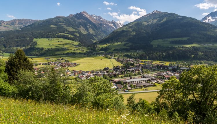 Ein kleines Dorf mit Feldern und Häusern liegt in einem grünen Tal, umgeben von hohen, schneebedeckten Bergen.