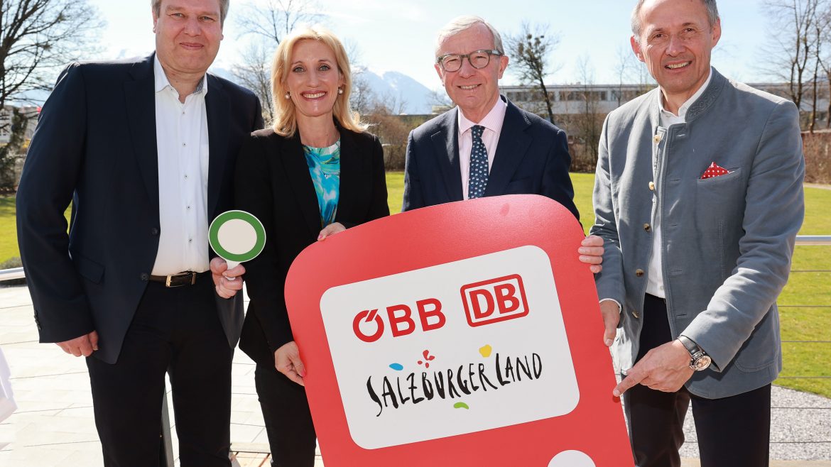 Vier lächelnde Menschen halten an einem sonnigen Tag im Freien ein rotes Salzburger Bahnschild.