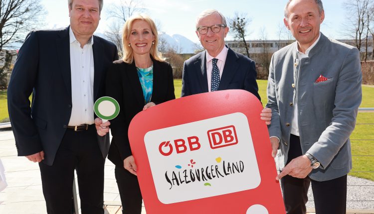Vier lächelnde Menschen halten an einem sonnigen Tag im Freien ein rotes Salzburger Bahnschild.