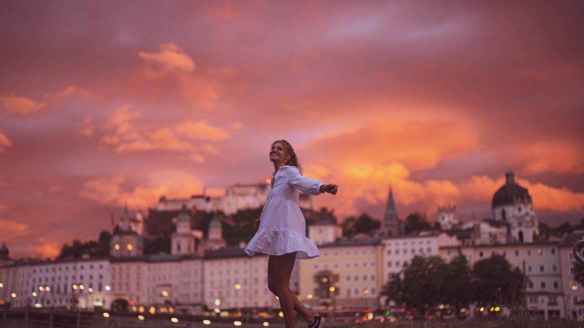 Eine Frau in einem weißen Kleid wirbelt fröhlich bei Sonnenuntergang, mit einer Stadt und einem dramatischen orangefarbenen Himmel im Hintergrund.