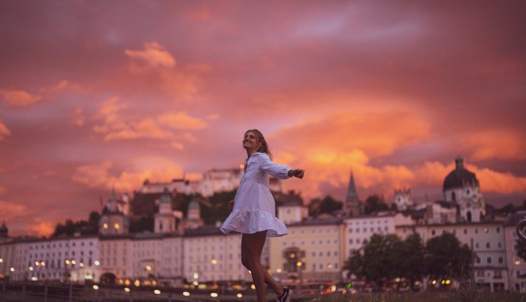 Eine Frau in einem weißen Kleid wirbelt fröhlich bei Sonnenuntergang, mit einer Stadt und einem dramatischen orangefarbenen Himmel im Hintergrund.