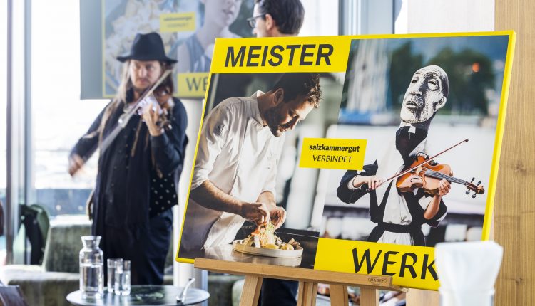 Ein Mann spielt Geige neben einem Plakat, das einen Koch und eine Geigenpuppe zeigt, mit dem Text MEISTER WERK.