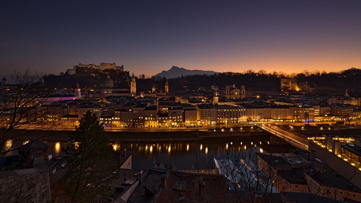 Eine Stadtlandschaft in der Abenddämmerung mit beleuchteten Gebäuden, einem Fluss und Bergen im Hintergrund.