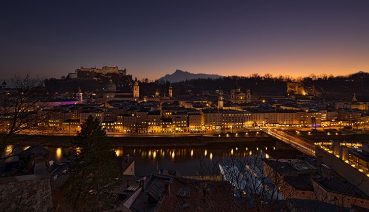 Eine Stadtlandschaft in der Abenddämmerung mit beleuchteten Gebäuden, einem Fluss und Bergen im Hintergrund.