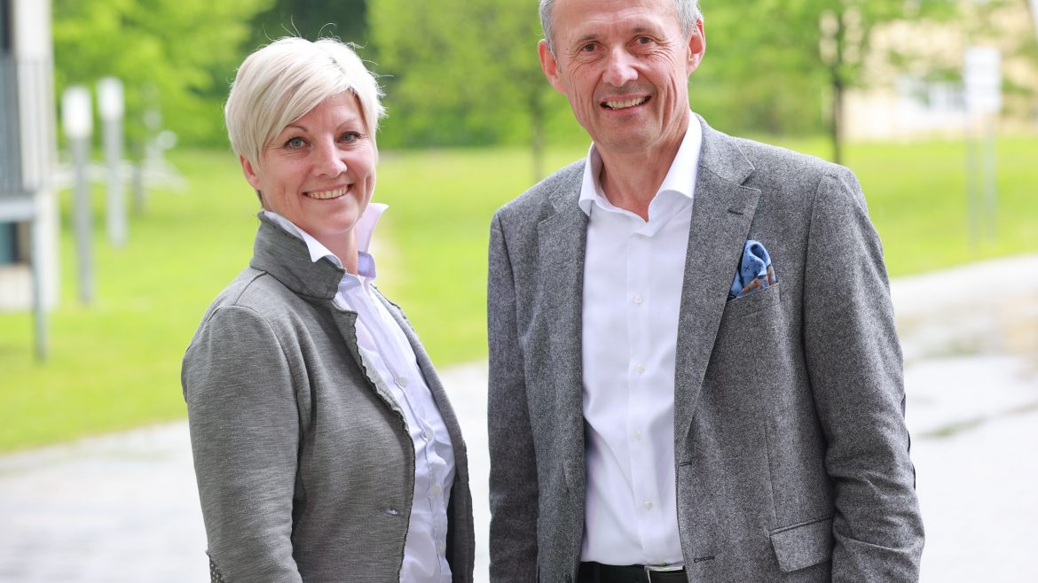 Zwei lächelnde Erwachsene in Geschäftskleidung stehen im Freien auf einem gepflasterten Weg mit grünen Bäumen im Hintergrund.