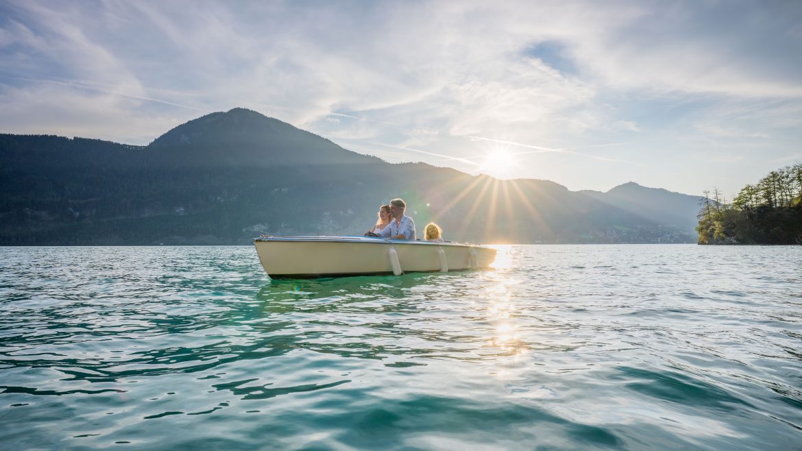Eine dreiköpfige Familie in einem Ruderboot auf einem See mit Bergen und der untergehenden Sonne im Hintergrund.