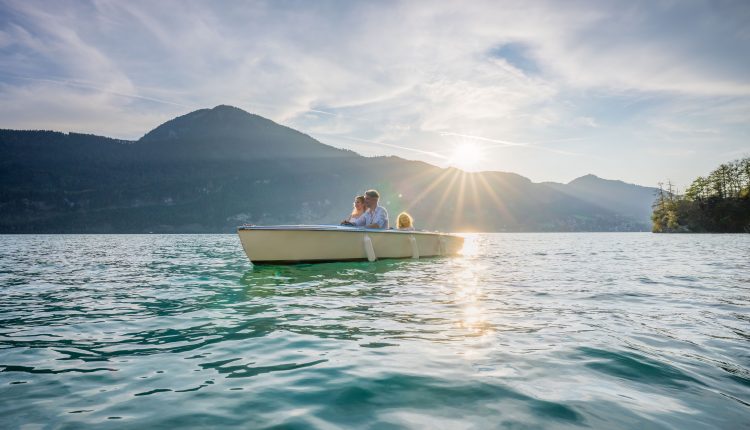 Eine dreiköpfige Familie in einem Ruderboot auf einem See mit Bergen und der untergehenden Sonne im Hintergrund.